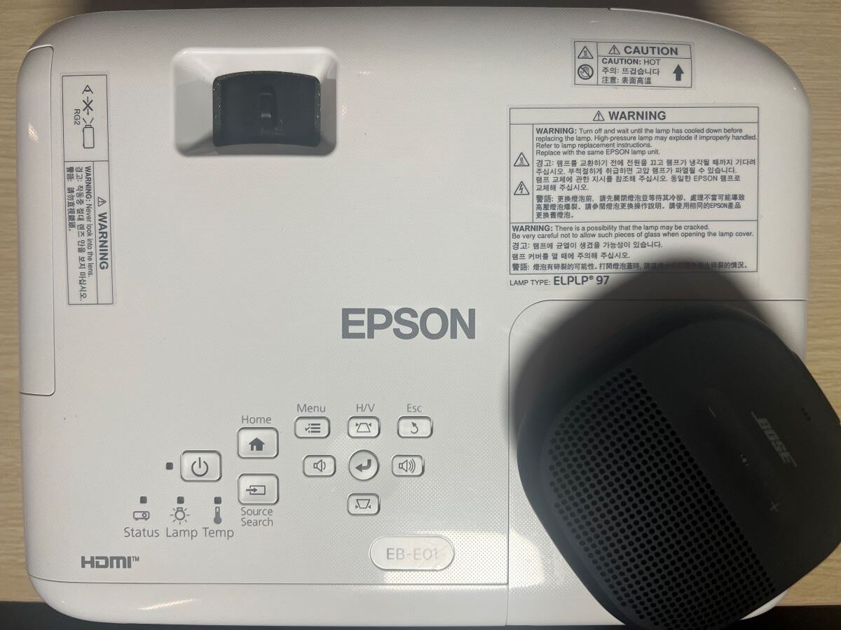 Bose speaker is lying on top of the Epson projector and both are on a wooden table