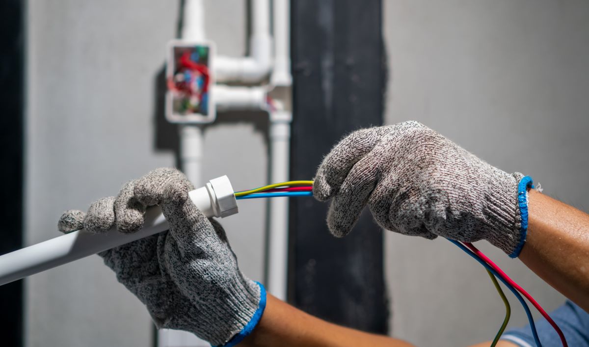 A hand wearing electrical gloves pushes the electrical wires through a white conduit