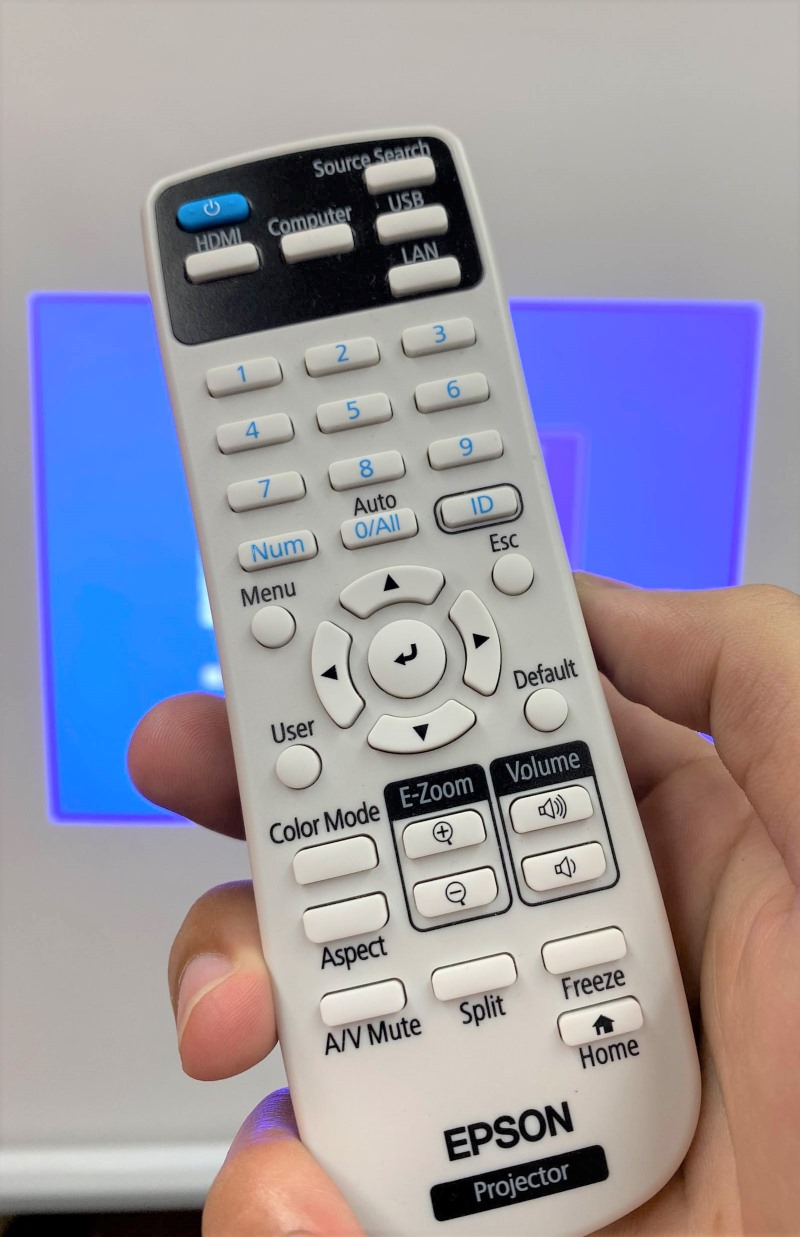 a hand holding an Epson projector control remote in front of the projector screen
