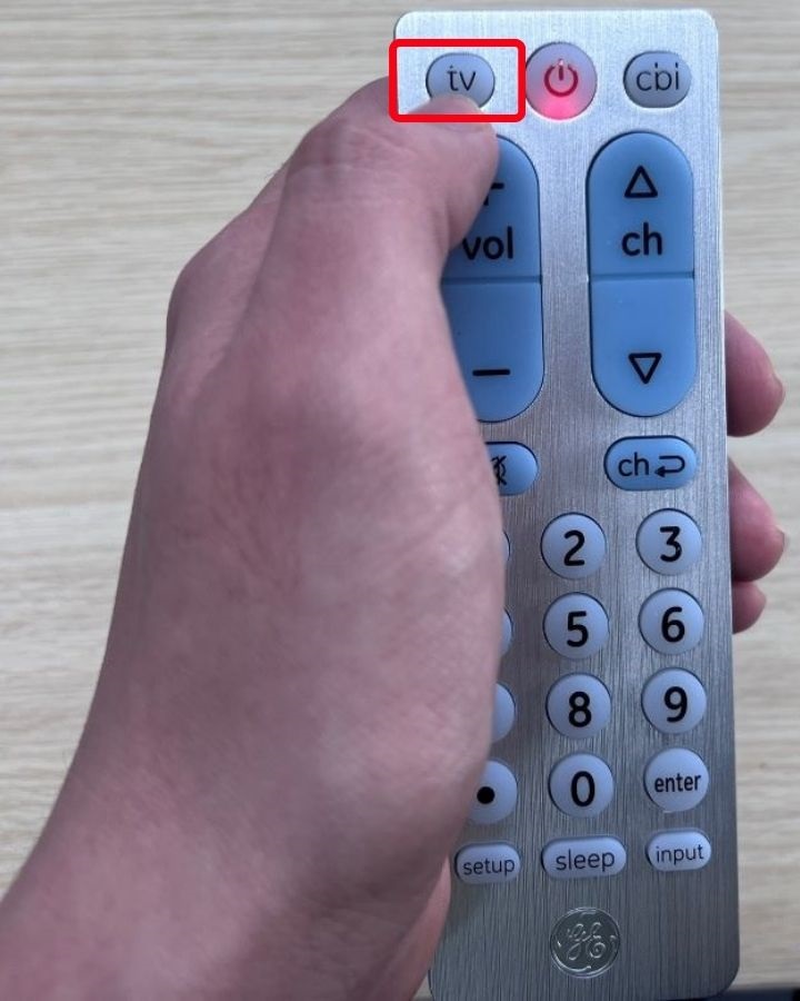 A hand is pressing the TV button on the GE universal remote