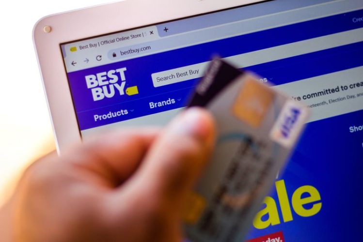 a person holds a bank card with Best Buy website shown in a laptop in the background