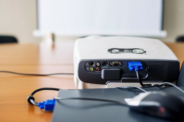 a white projector connect to laptop by a cable with blurr projector screen on the background