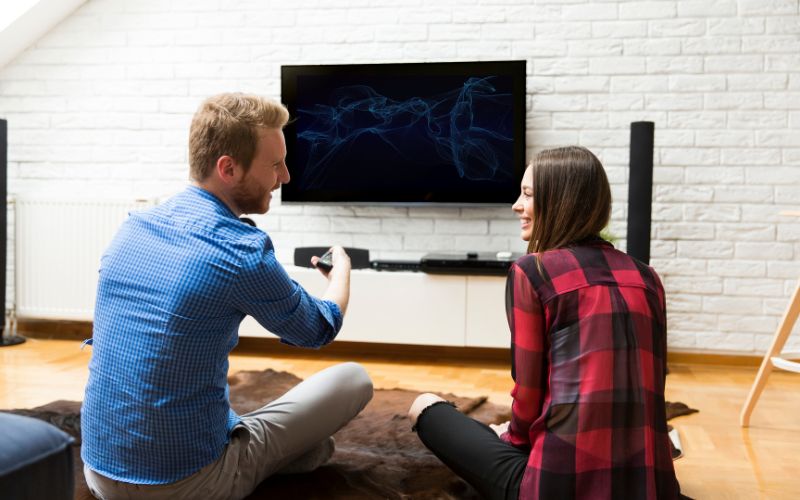man is holding a romote control while sitting comfortably on the floor and watching tv with his girlfriend