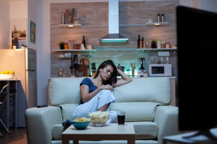 a woman tiredly falling asleep on sofa with the TV on in her apartment