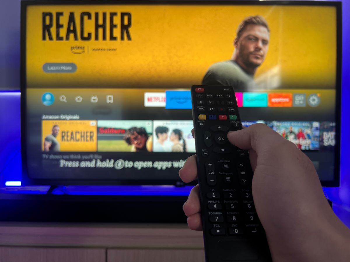 A hand is using a universal remote to control Fire TV which is showing on the TV screen