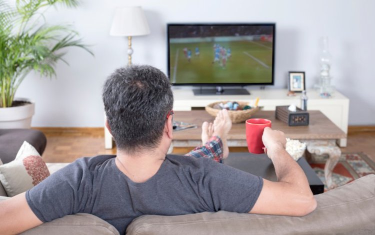 a man is sitting lazily on the couch watching football match on TV