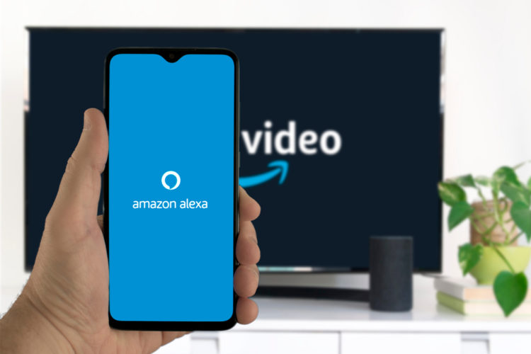a hand is holding a smartphone showing amazon alexa app's startup screen with a tv in the background