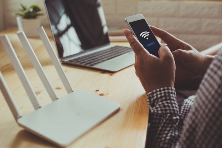 a router and a laptop is on the table, the man is holding a smartphone with his hands laying on the table, there is a wi-fi symbol on the smartphone