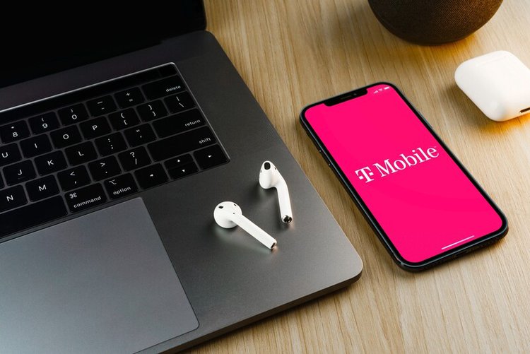 a phone with T-mobile app on the screen next to a laptop and airpod case
