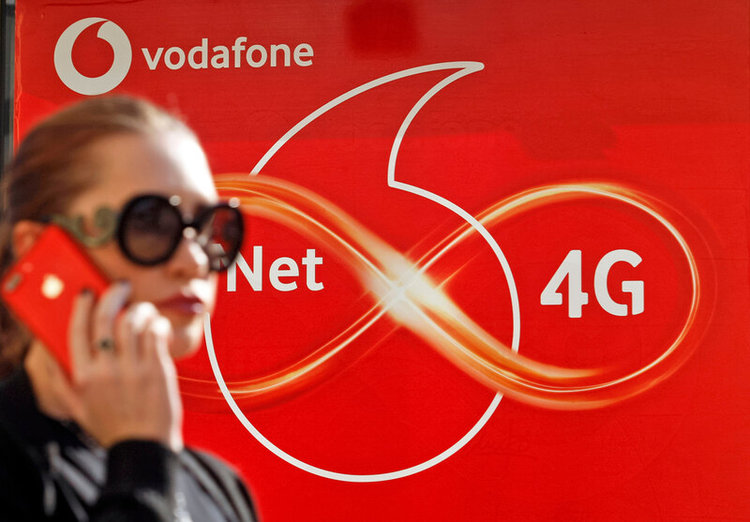 a woman in black glasses speaking on a phone with Vodafone logo in the background