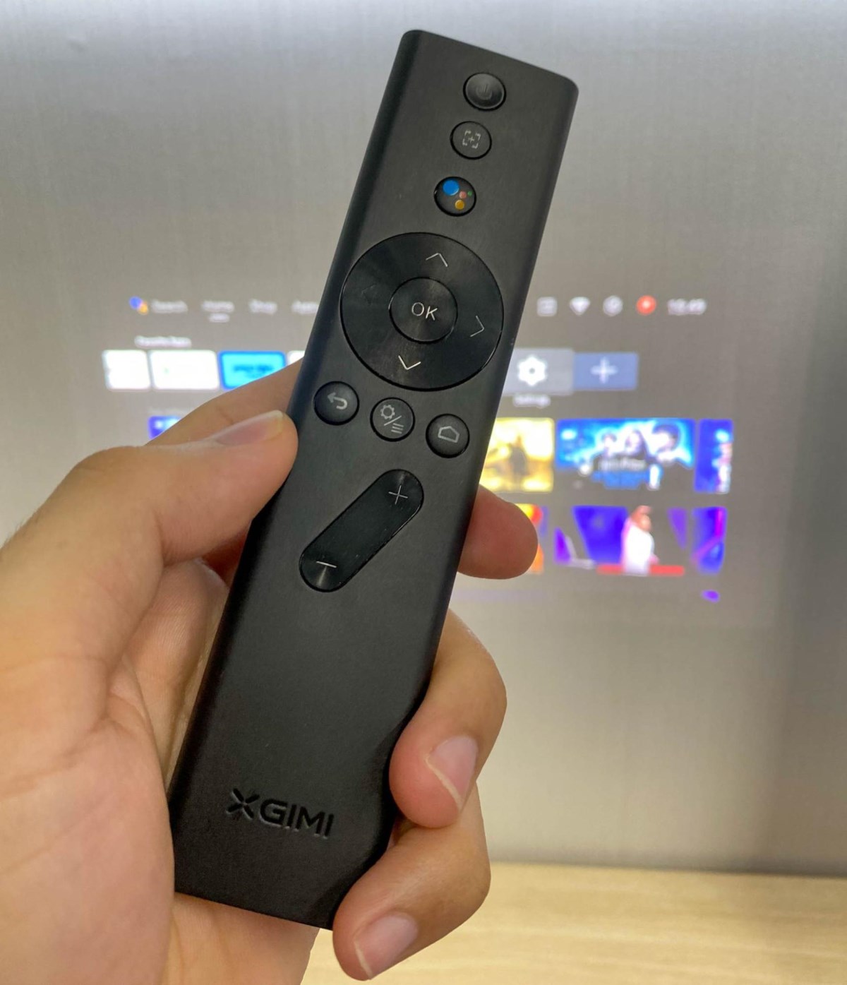 a hand is holding an XGIMI projector remote in front of the projector screen