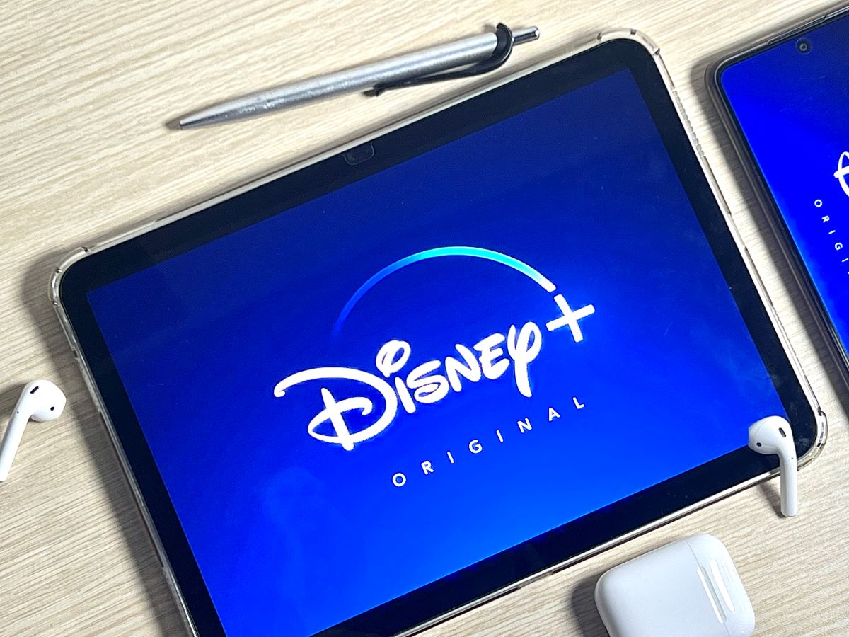 an ipad showing disney plus logo, with airpods, a pen, and a phone nearby