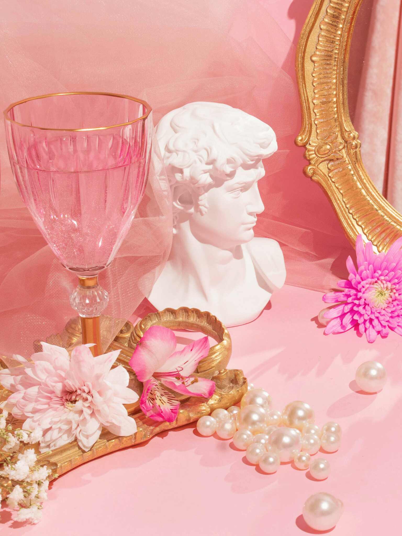 a white statue with flowers, pearls, a glass and a mirror on pink fabric
